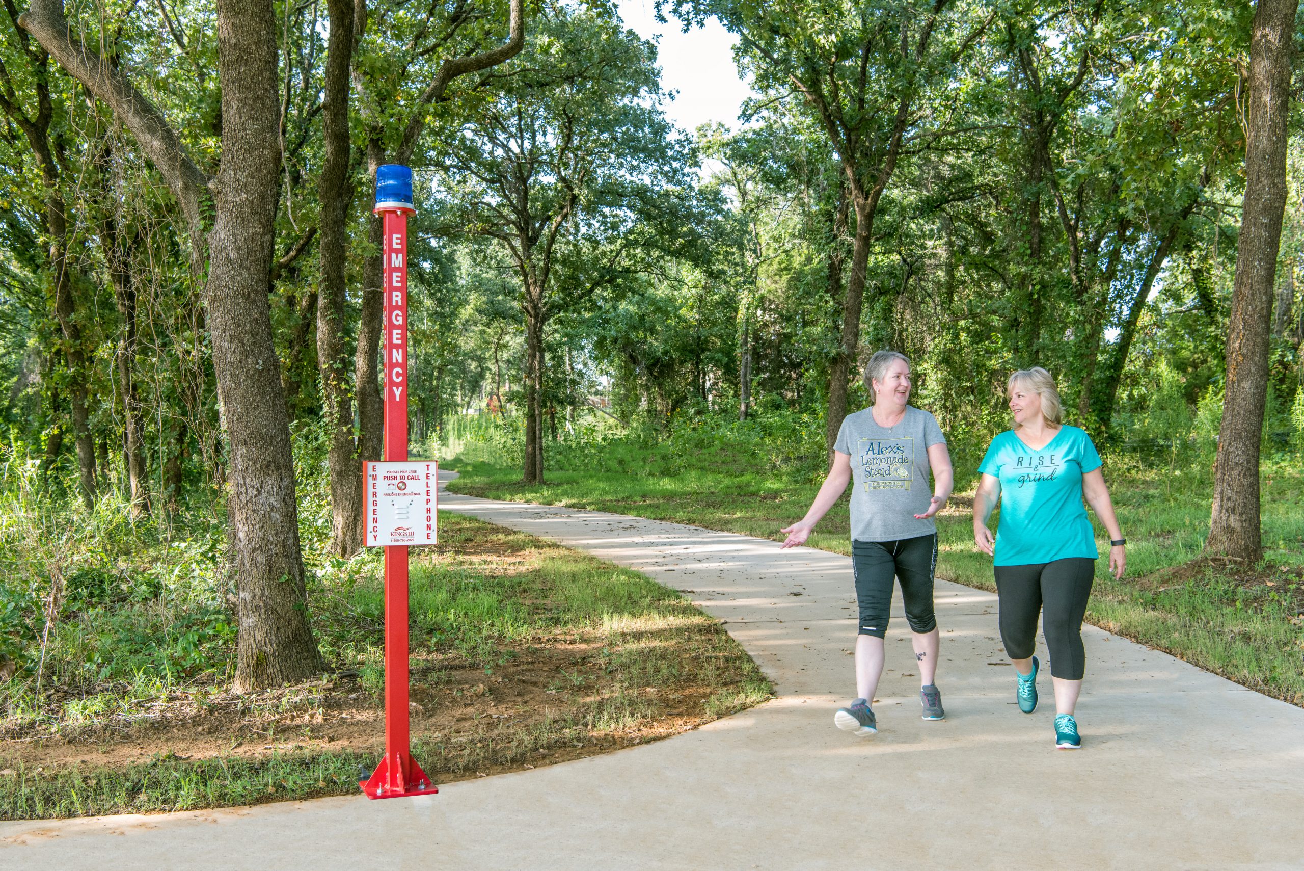 Two women walking on a path in the woods, surrounded by tall trees and lush greenery passing by a tall red emergency help phone